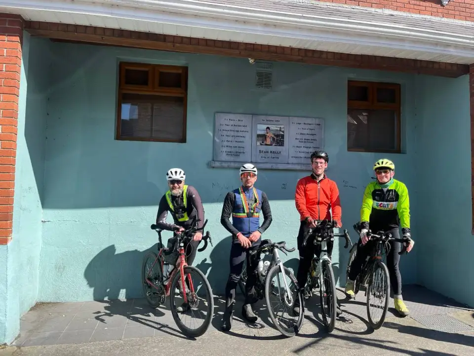 Four randonneurs with their bikes under the Sean Kelly plaque in Carrick-on-Suir