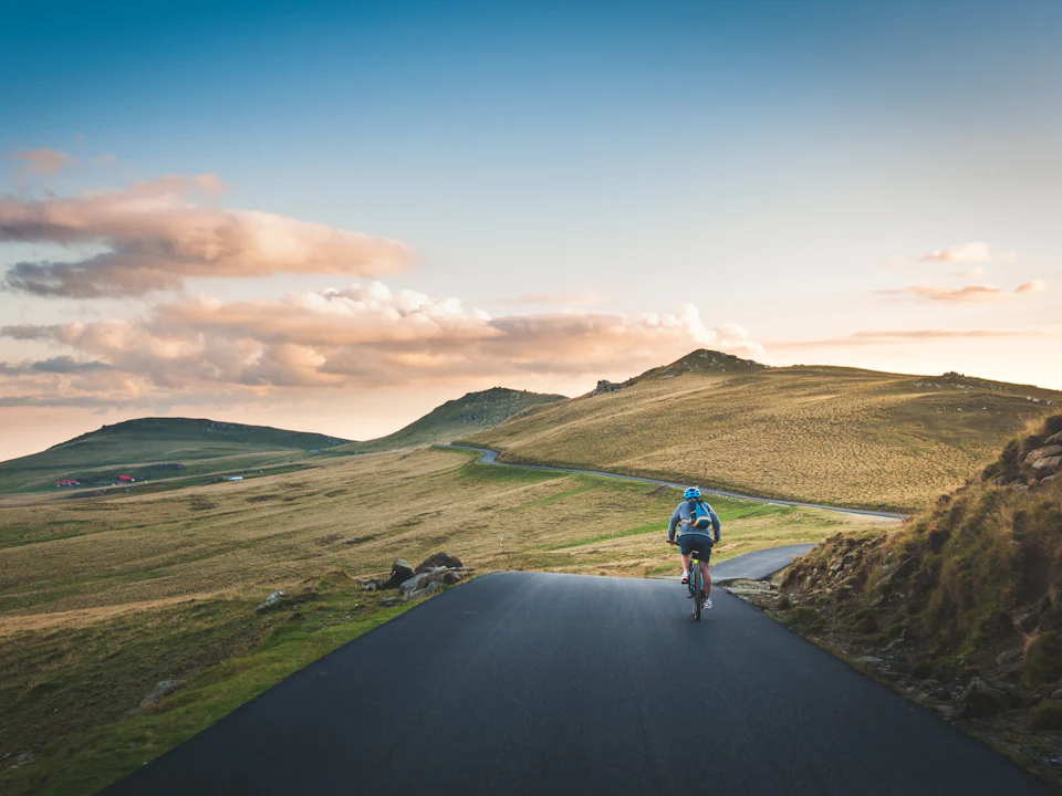 A cyclist on an empty road heading towards green hills in evening light