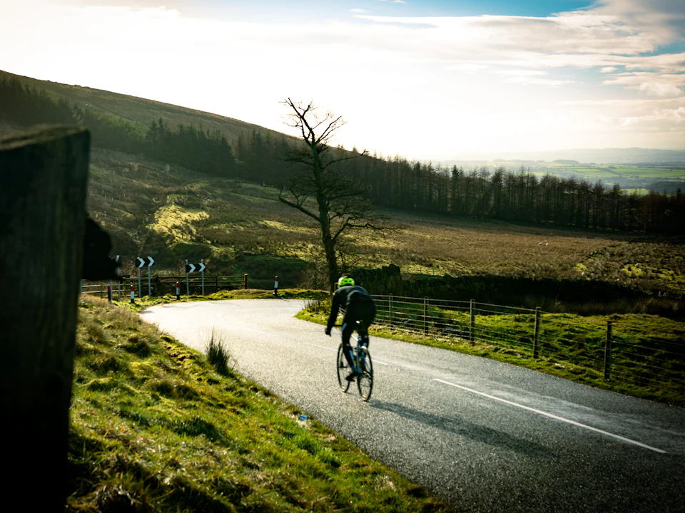 A cyclist climbing a winding upland road past forestry in low winter sun