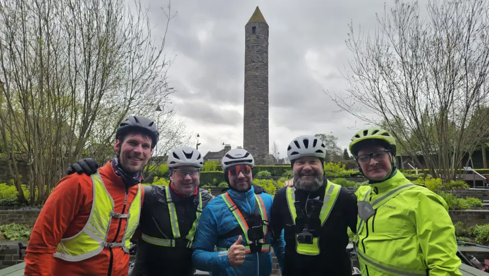 Five riders in hi-vis standing with their bikes in front of an Irish round tower