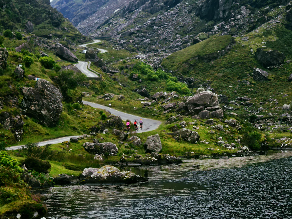 Cyclists on a narrow road winding through a rocky mountain pass above a lake
