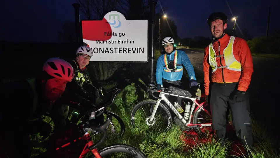 Four randonneurs with lit bikes at the Monasterevin town sign at night
