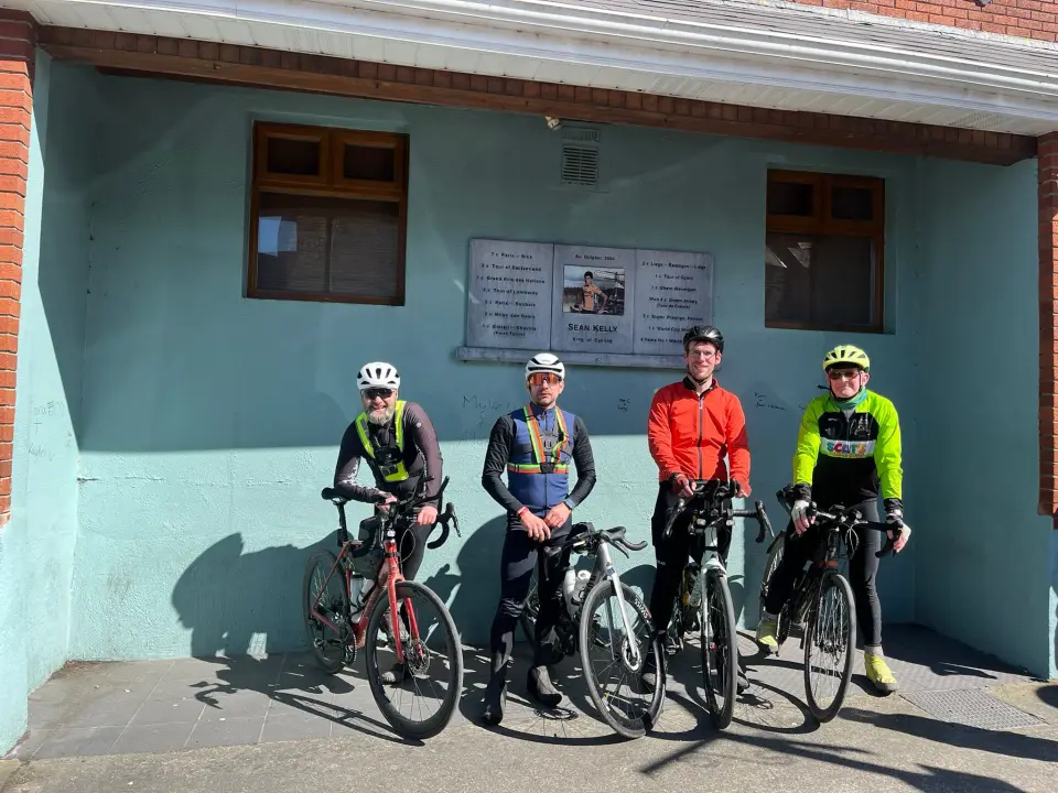 Four randonneurs standing with their bikes in front of a plaque honouring Sean Kelly